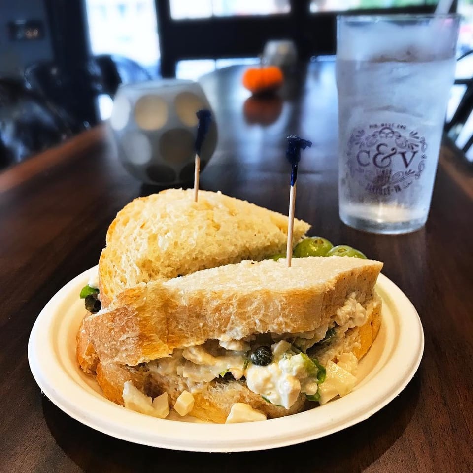 Hearty deli sandwich on crusty bread with creamy filling, pickles, and potato salad on a plate beside a branded water glass.