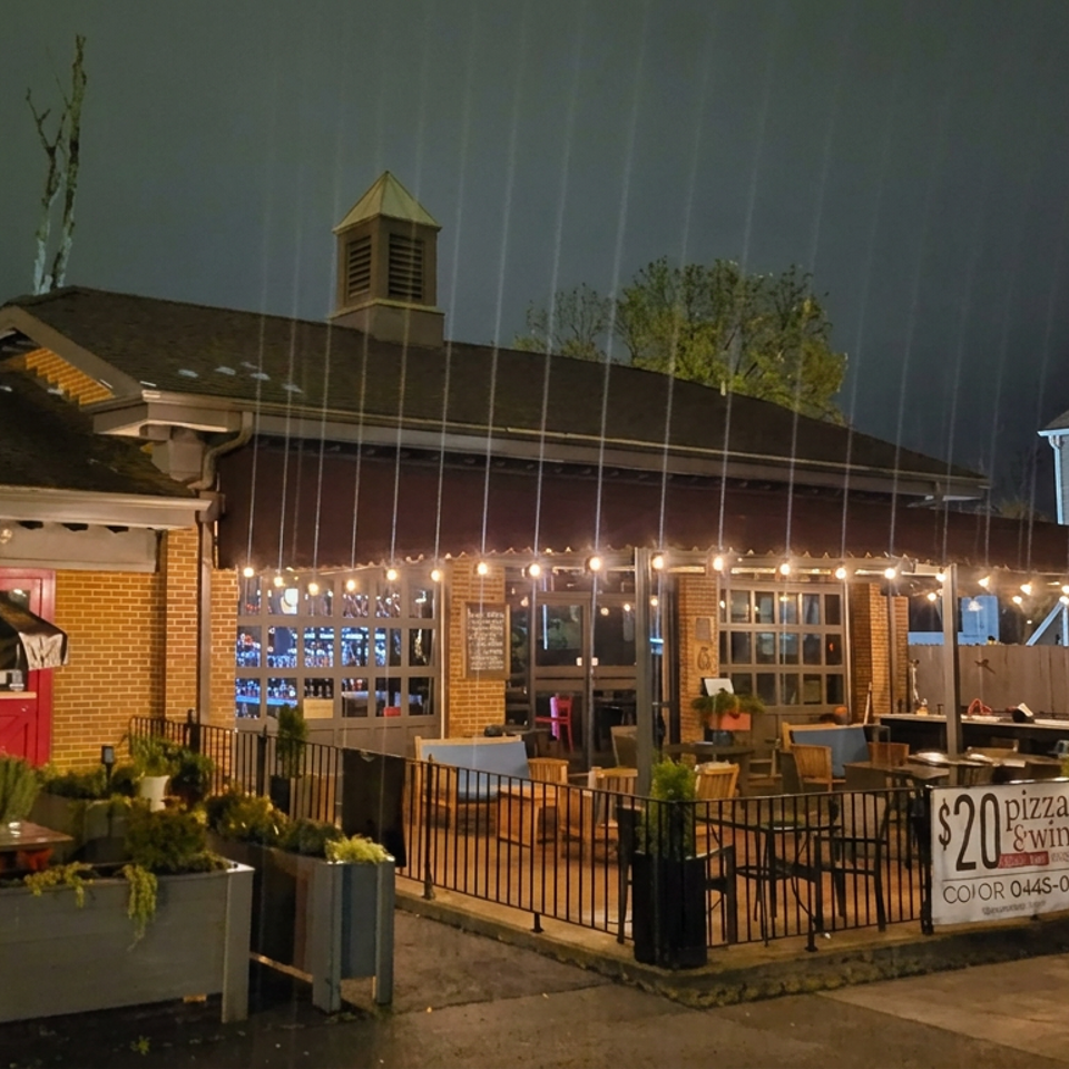 Rainy evening view of a brick pizza restaurant patio with string lights, outdoor seating, planters, and a $20 pizza and wine sign.