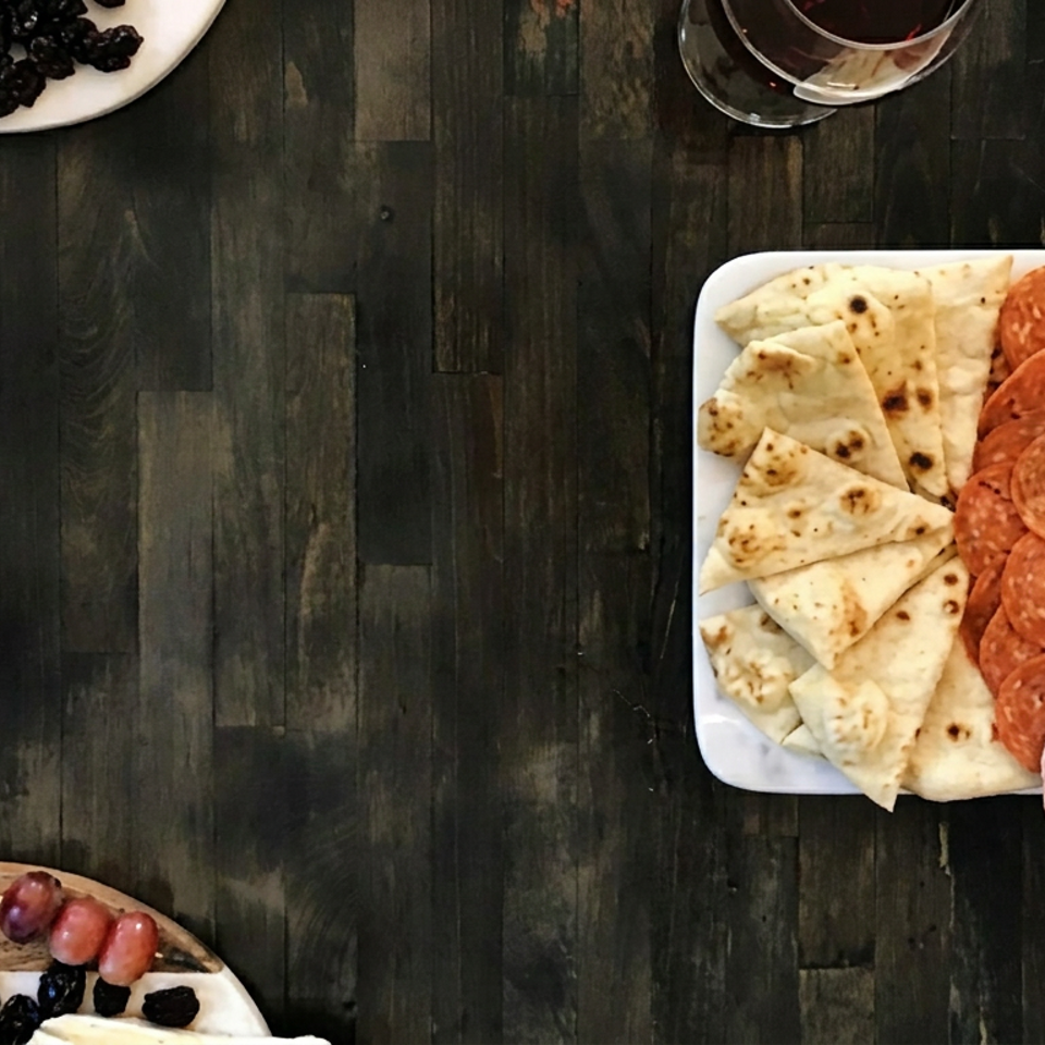 Overhead spread of flatbread wedges, pepperoni, rolled ham, olives, and skewered appetizers on a dark wooden table.
